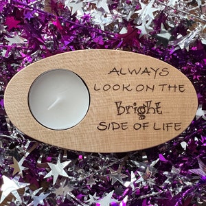 May include: Wooden tea light holder with a round cutout for a candle. The holder has the text "Always look on the bright side of life" engraved on it.