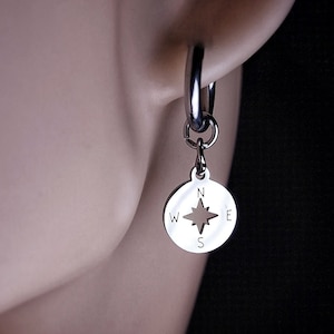 May include: Silver hoop earrings with a compass charm. The compass charm has a cut-out design of a compass rose with the letters N, S, E, and W.