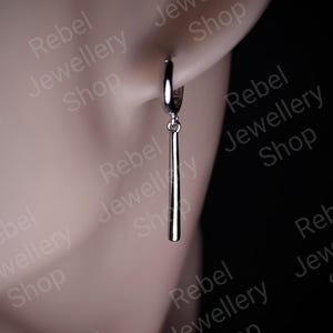 May include: A single silver earring featuring a long, thin bar pendant suspended from a hoop. The earring is attached to an ear. The background includes the text "Rebel Jewellery Shop".
