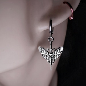 Könnte beinhalten: Silberne Creolen mit einem Anhänger in Form eines Totenkopfschwärmers mit einem Schädel und einem Dolch. Der Totenkopfschwärmer ist detailliert mit Flügeln und einem Körper.