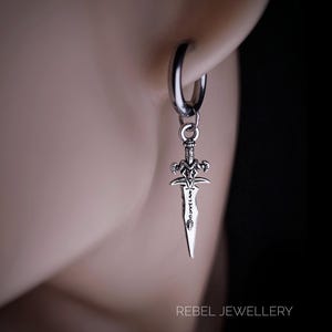 May include: A silver-coloured earring with a small dagger pendant. The dagger has detailed features on the hilt and blade. The earring is a hoop style, and the pendant hangs freely. The text "REBEL JEWELLERY" is visible.