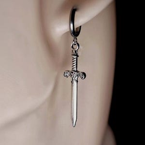 May include: A silver-colored sword charm earring hanging from a black hoop earring. The sword has a detailed hilt and blade. The earring is shown on a person's ear. The text "REBEL JEWELLERY" is at the bottom.