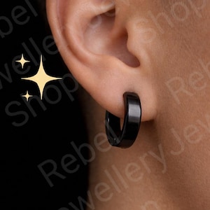 May include: A close-up of a black hoop earring on an ear. The earring is a simple, solid black color and has a smooth, polished finish. The earring is a classic style, suitable for everyday wear.