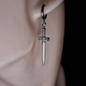 Könnte beinhalten: Ein silberfarbener Creolen-Ohrring mit einem hängenden Schwertanhänger. Das Schwert hat ein detailliertes Heft mit komplexen Verzierungen und eine gerade, spitze Klinge. Der Text REBEL JEWELLERY ist in der unteren rechten Ecke zu sehen.