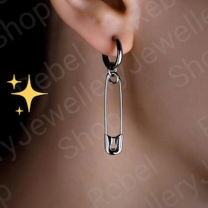 May include: A silver safety pin earring hanging from a black hoop earring. The earring is shown worn on an ear. The background is blurred, focusing attention on the jewellery.