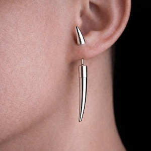 May include: A silver-colored earring set featuring a small stud and a long, pointed dangle. The earrings are worn on the ear, with the dangle extending downwards. The background is black, highlighting the jewelry.
