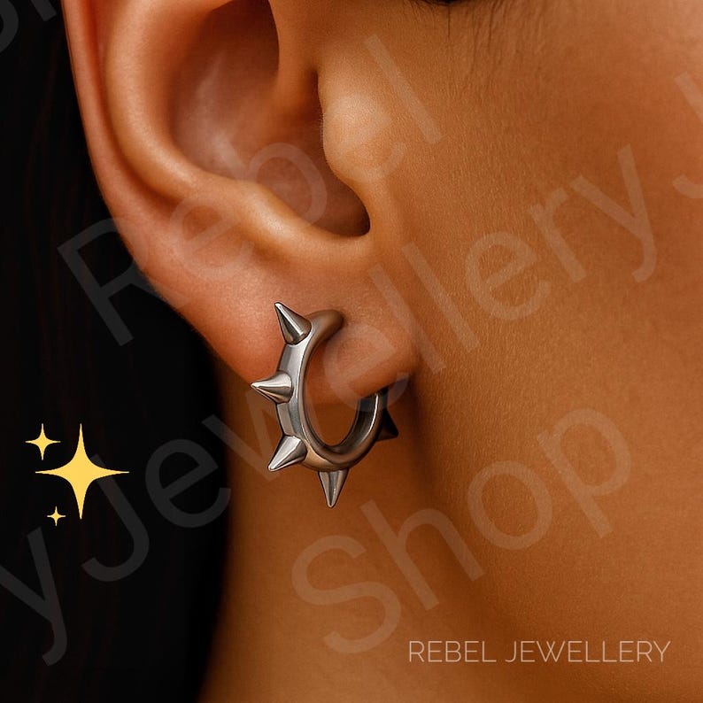 May include: A silver-toned hoop earring featuring four prominent, evenly spaced conical spikes along the outer edge. The earring is secured through the earlobe, showcasing a metallic finish and a circular design with sharp, pointed protrusions.