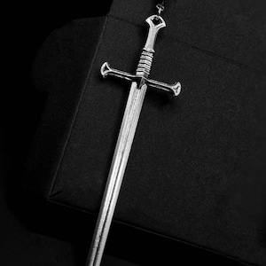 May include: A silver sword pendant with a detailed design. The pendant is hanging from a black background with the text 'REBEL JEWELLERY' at the bottom.