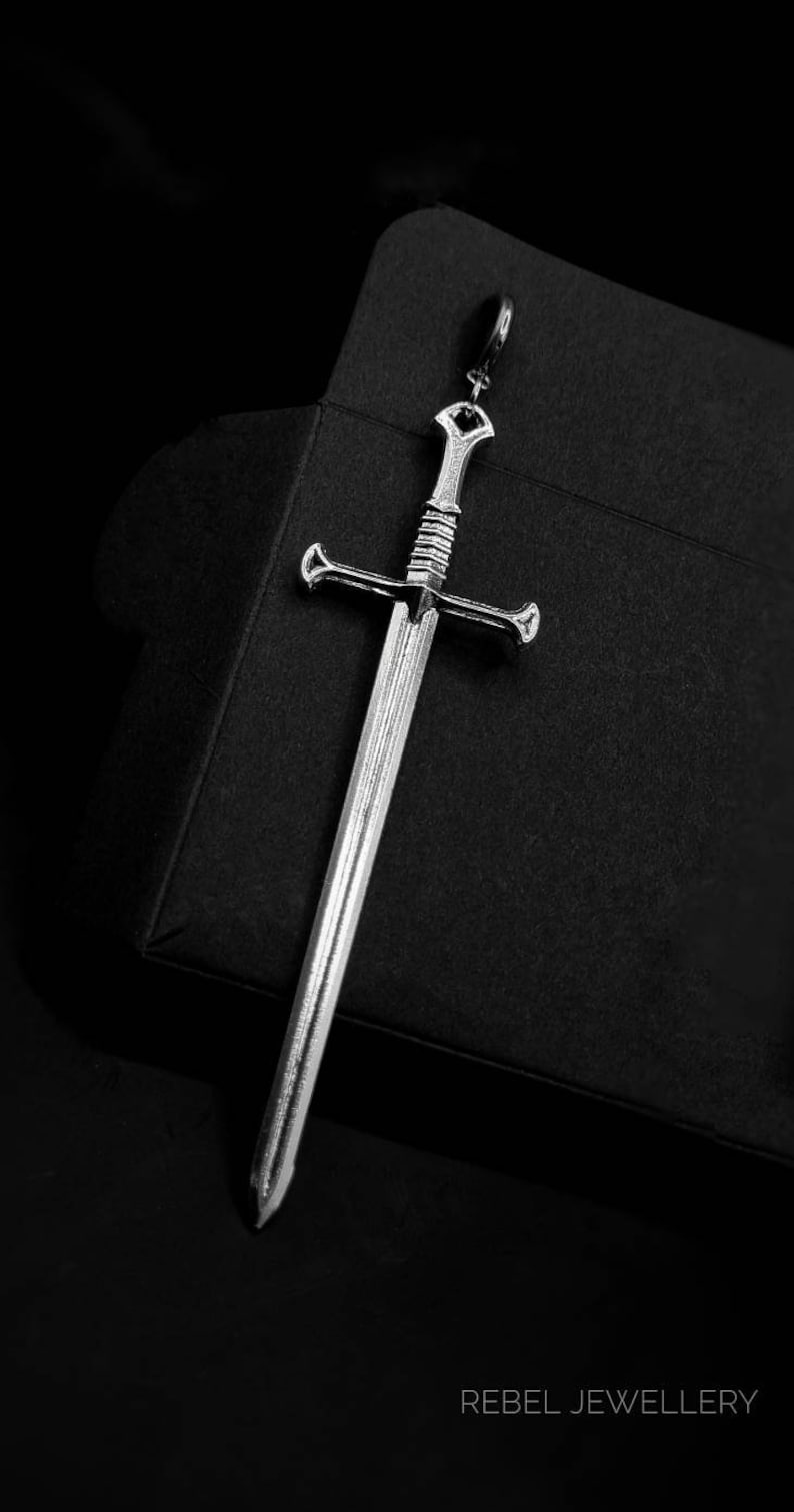May include: A silver sword pendant with a detailed design. The pendant is hanging from a black background with the text 'REBEL JEWELLERY' at the bottom.