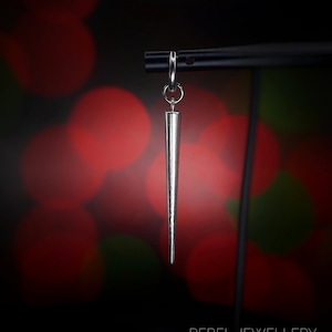 May include: A silver-colored, cone-shaped earring suspended from a black metal bar. The earring has a pointed tip and a small hoop at the top. The background is a blurred mix of red and green lights. The text "REBEL JEWELLERY" is at the bottom.