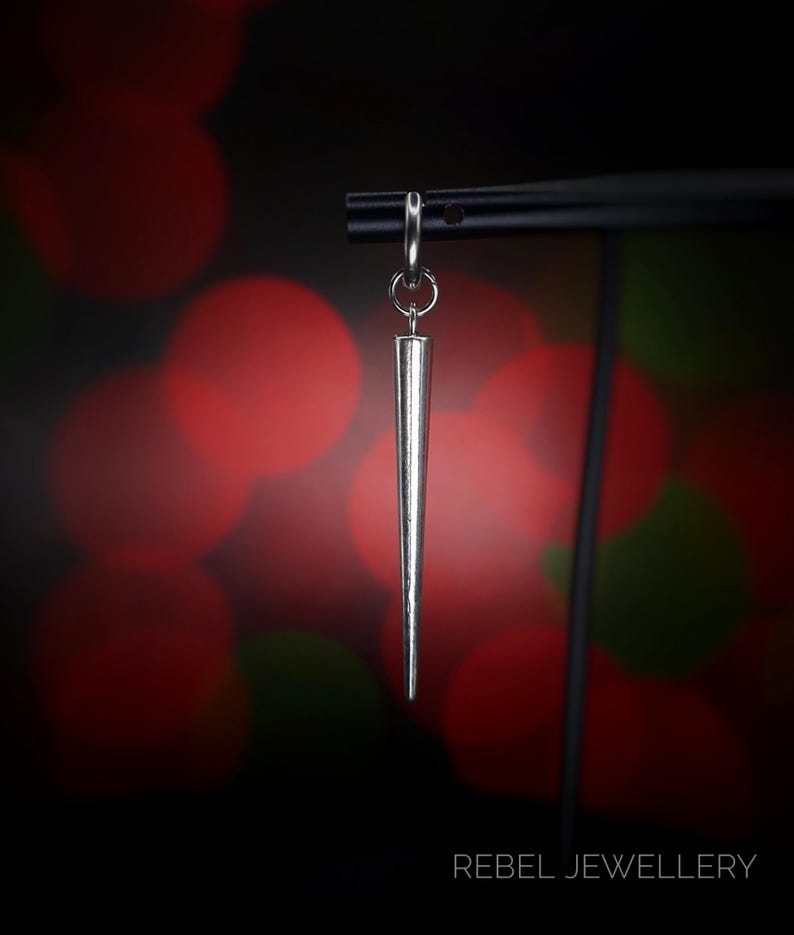 May include: A silver-colored, cone-shaped earring suspended from a black metal bar. The earring has a pointed tip and a small hoop at the top. The background is a blurred mix of red and green lights. The text "REBEL JEWELLERY" is at the bottom.