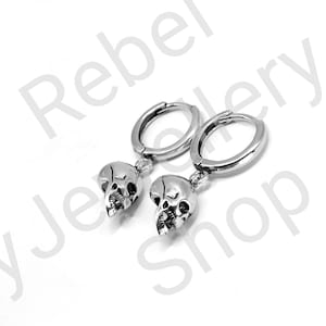 May include: A pair of silver-colored hoop earrings. Each earring features a small skull pendant. The earrings are displayed against a white background. The text "Rebel Jewelry Shop" is visible in the background.