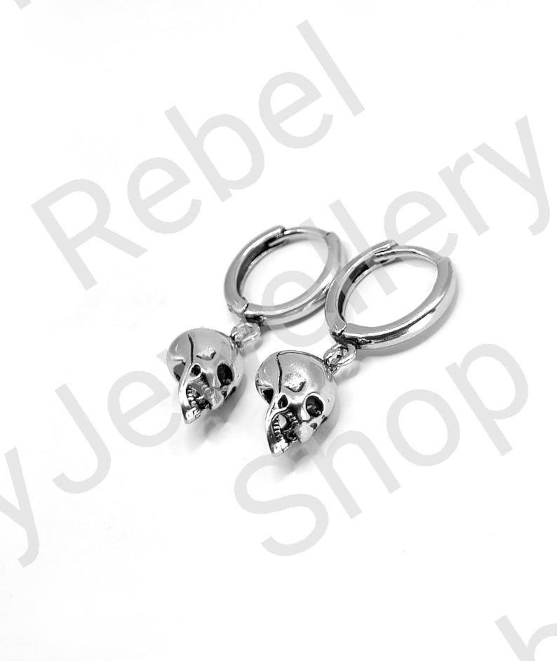 May include: A pair of silver-colored hoop earrings. Each earring features a small skull pendant. The earrings are displayed against a white background. The text "Rebel Jewelry Shop" is visible in the background.