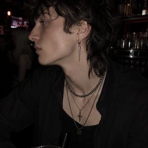 May include: A person wearing a black shirt, layered necklaces, and a spike earring. The person has dark hair and a nose ring. The background is dark, with a bar setting.