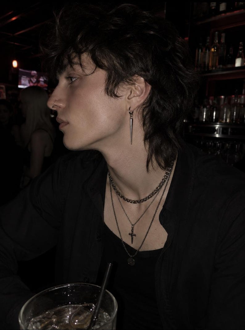 May include: A person wearing a black shirt, layered necklaces, and a spike earring. The person has dark hair and a nose ring. The background is dark, with a bar setting.