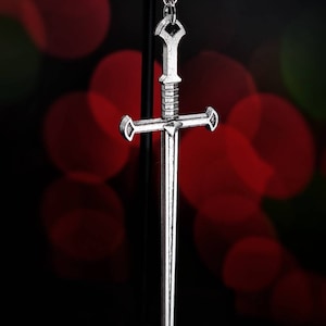 May include: A silver sword pendant with a cross-shaped design. The pendant is hanging from a black stand against a red and green blurred background. The text 'REBEL JEWELLERY' is visible at the bottom of the image.