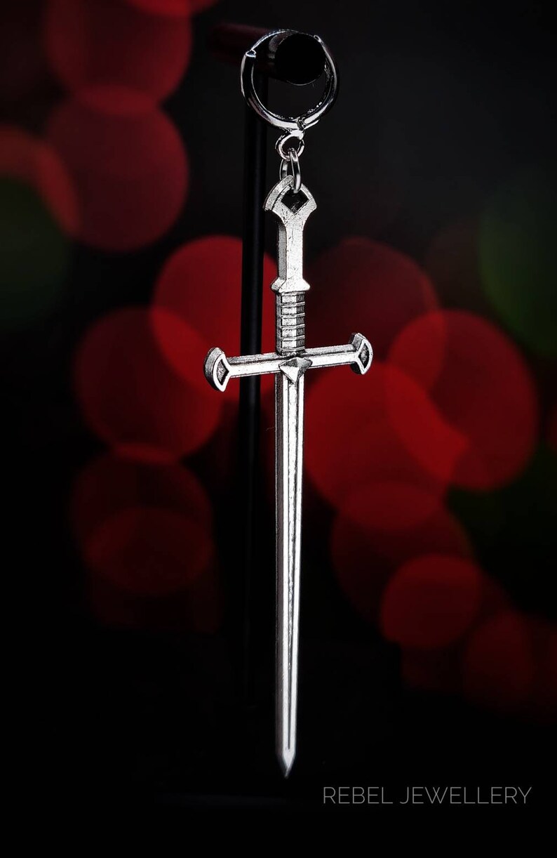 May include: A silver sword pendant with a cross-shaped design. The pendant is hanging from a black stand against a red and green blurred background. The text 'REBEL JEWELLERY' is visible at the bottom of the image.