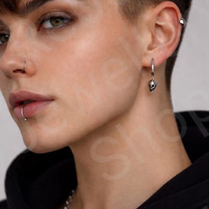 May include: A silver hoop earring with a small skull charm. The earring is worn on the ear of a person with short hair and a nose ring. A silver chain necklace is also visible.