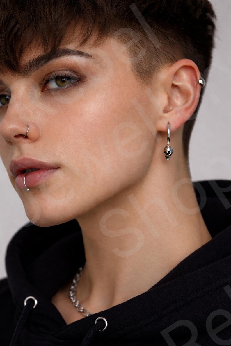 May include: A silver hoop earring with a small skull charm. The earring is worn on the ear of a person with short hair and a nose ring. A silver chain necklace is also visible.
