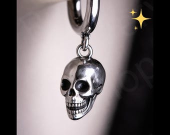 May include: A silver-colored hoop earring with a skull charm. The skull has detailed features, including eye sockets, a nose cavity, and teeth. The earring is suspended from a silver hoop, creating a gothic or alternative aesthetic.