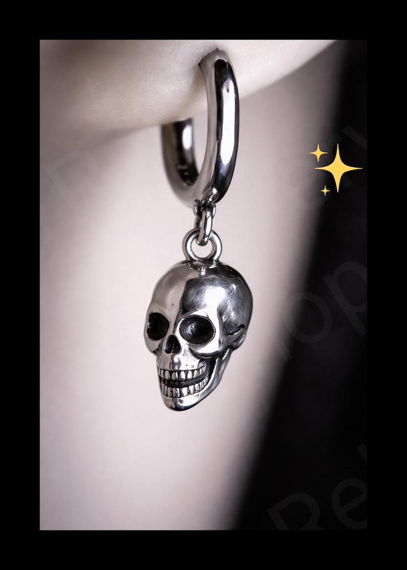 May include: A silver-colored hoop earring with a skull charm. The skull has detailed features, including eye sockets, a nose cavity, and teeth. The earring is suspended from a silver hoop, creating a gothic or alternative aesthetic.