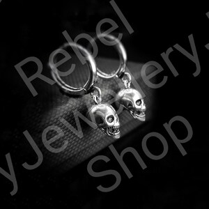May include: A pair of silver-colored skull earrings. Each earring features a detailed skull pendant hanging from a hoop. The skulls have visible teeth and eye sockets. The background is dark, emphasizing the jewelry.