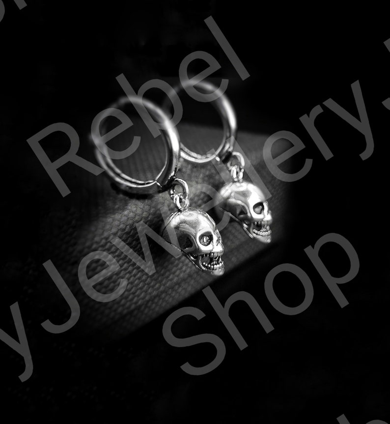 May include: A pair of silver-colored skull earrings. Each earring features a detailed skull pendant hanging from a hoop. The skulls have visible teeth and eye sockets. The background is dark, emphasizing the jewelry.