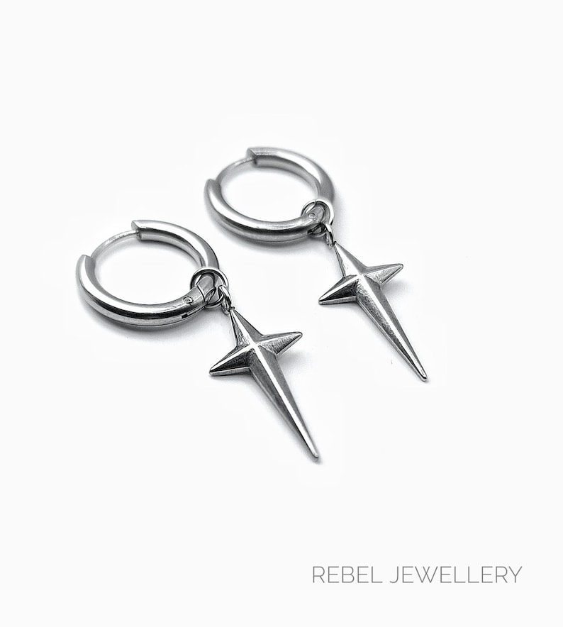May include: A pair of silver-colored hoop earrings with star-shaped pendants. The earrings are made of a shiny metal and hang from the hoops. The text "REBEL JEWELLERY" is at the bottom of the image.