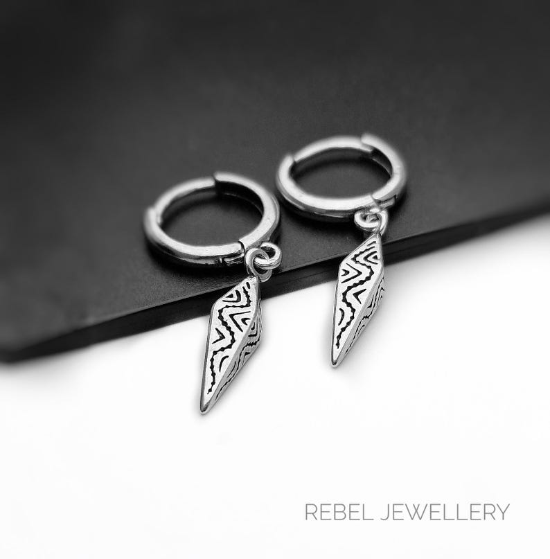 May include: A pair of silver hoop earrings with dangling, diamond-shaped pendants. Each pendant features a black and white geometric pattern. The earrings are displayed on a dark surface, with the text "REBEL JEWELLERY" visible in the bottom right corner.