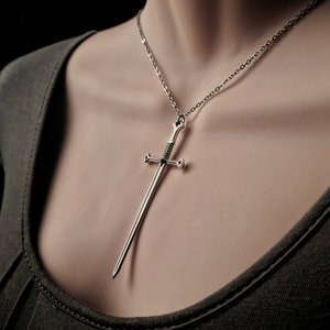 May include: A silver sword pendant necklace on a silver chain. The sword has a detailed design and is hanging on a mannequin's neck.