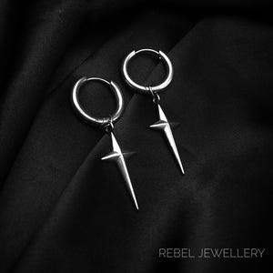 May include: A pair of silver hoop earrings with a starburst design. The earrings are suspended from the hoops and have a pointed, star-shaped pendant. The earrings are displayed on a black fabric background. The text "REBEL JEWELLERY" is at the bottom.