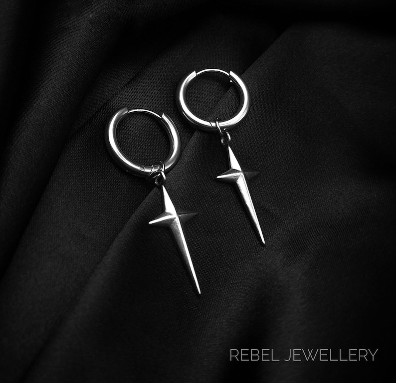 May include: A pair of silver hoop earrings with a starburst design. The earrings are suspended from the hoops and have a pointed, star-shaped pendant. The earrings are displayed on a black fabric background. The text "REBEL JEWELLERY" is at the bottom.