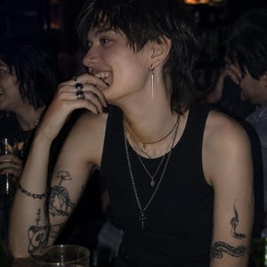 May include: A person wearing a black tank top, multiple silver necklaces, and a silver earring. The person has tattoos on their arms and is smiling. A glass of liquid with a lemon slice is on the table.