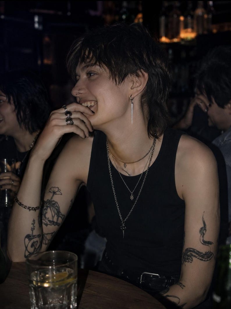 May include: A person wearing a black tank top, multiple silver necklaces, and a silver earring. The person has tattoos on their arms and is smiling. A glass of liquid with a lemon slice is on the table.