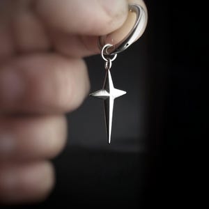 May include: A silver-colored earring with a star pendant. The star has four points and hangs from a hoop. The earring is held against a dark background. The text "REBEL JEWELLERY" is at the bottom.