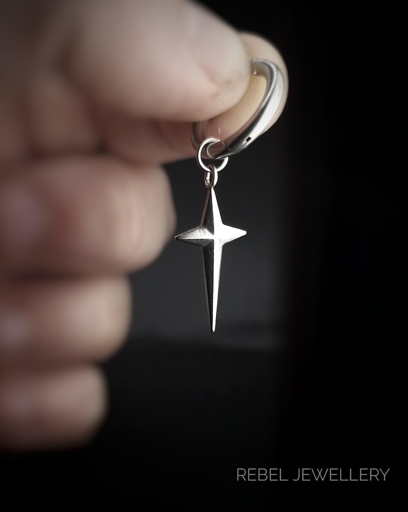 May include: A silver-colored earring with a star pendant. The star has four points and hangs from a hoop. The earring is held against a dark background. The text "REBEL JEWELLERY" is at the bottom.