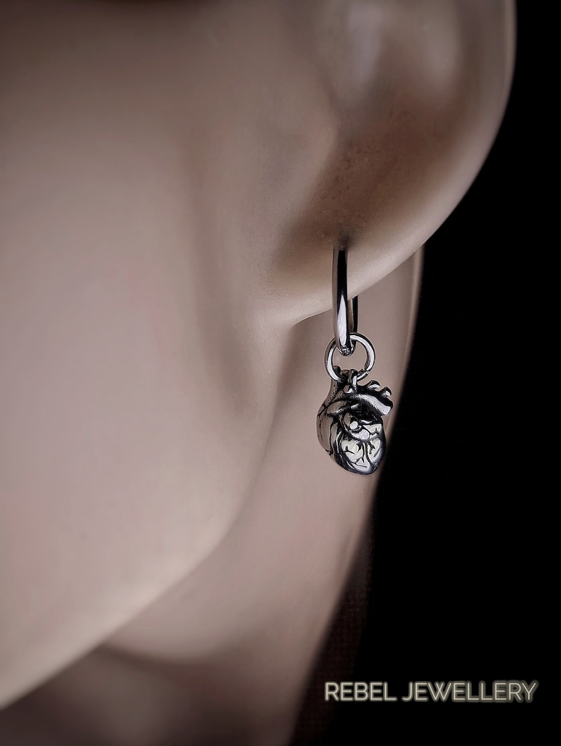 May include: A silver hoop earring featuring a detailed anatomical heart pendant. The earring is attached to an ear, with the text "REBEL JEWELLERY" visible in the lower right corner.