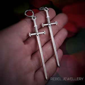 May include: Two silver sword earrings. The swords have a cross guard and a pointed tip. The earrings are hanging from a hand.