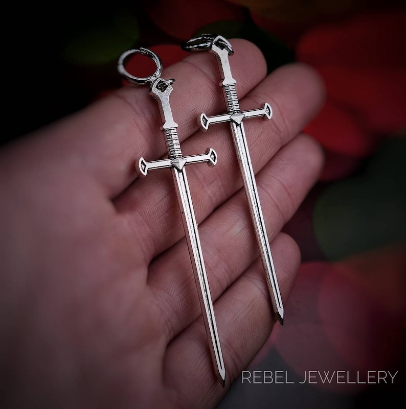 May include: Two silver sword earrings. The swords have a cross guard and a pointed tip. The earrings are hanging from a hand.