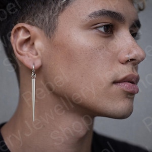 May include: A silver-colored, long, pointed earring hangs from a silver hoop. The earring is a simple, minimalist design. The earring is worn on the left ear. The person in the image is wearing a black shirt.