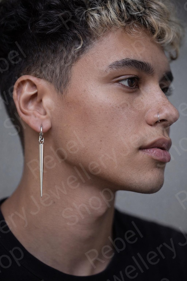 May include: A silver-colored, long, pointed earring hangs from a silver hoop. The earring is a simple, minimalist design. The earring is worn on the left ear. The person in the image is wearing a black shirt.