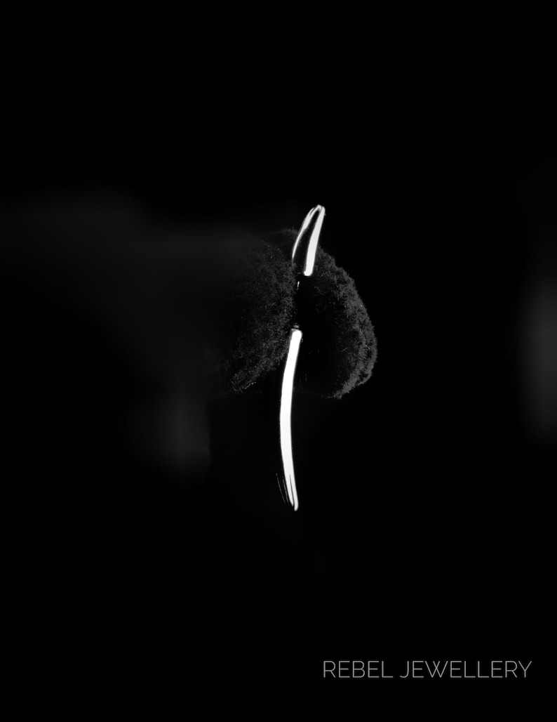 Puede incluir: Un pendiente plateado minimalista con un dise&ntilde;o curvo, sobre un fondo oscuro. El pendiente es una sola l&iacute;nea elegante, que refleja la luz. El texto "REBEL JEWELLERY" est&aacute; en la parte inferior.