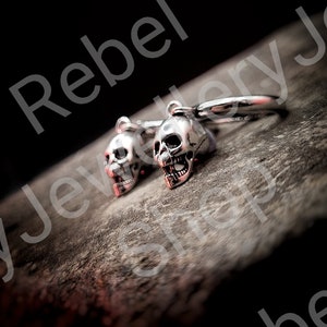 May include: Silver skull earrings with red accents. The earrings are detailed with open mouths and visible teeth. The image is set against a dark background, highlighting the jewelry's design. The earrings are a unique accessory.