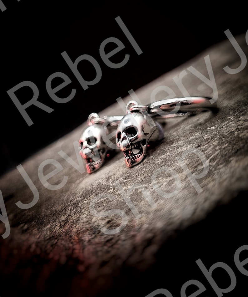 May include: Silver skull earrings with red accents. The earrings are detailed with open mouths and visible teeth. The image is set against a dark background, highlighting the jewelry's design. The earrings are a unique accessory.