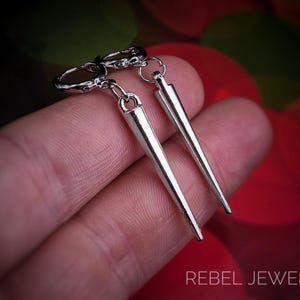 Puede incluir: Pendientes de punta plateados con un dise&ntilde;o puntiagudo. Los pendientes cuelgan de un peque&ntilde;o aro y se sostienen en una mano. El texto "REBEL JEWELLERY" es visible en la parte inferior de la imagen.