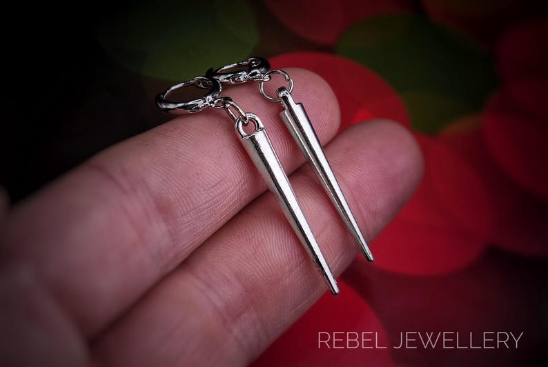 Puede incluir: Pendientes de punta plateados con un dise&ntilde;o puntiagudo. Los pendientes cuelgan de un peque&ntilde;o aro y se sostienen en una mano. El texto "REBEL JEWELLERY" es visible en la parte inferior de la imagen.