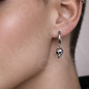 May include: Silver hoop earring with a small skull charm. The skull has detailed features and hangs from a simple, polished silver hoop. The earring is worn on the ear.
