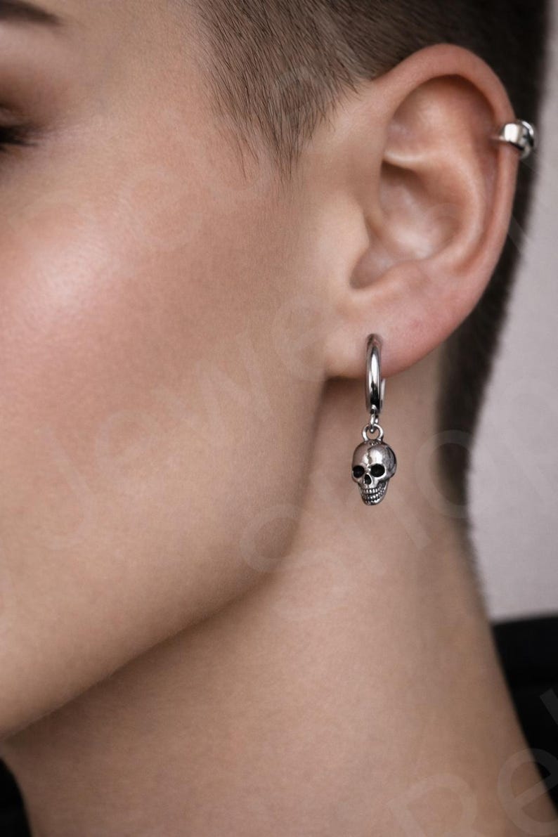 May include: Silver hoop earring with a small skull charm. The skull has detailed features and hangs from a simple, polished silver hoop. The earring is worn on the ear.