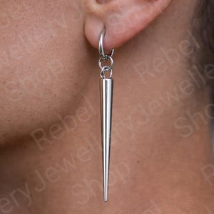 May include: A silver-colored, pointed earring is suspended from a small hoop. The earring has a long, conical shape, tapering to a sharp point. The jewelry is worn on the ear. The background is blurred.