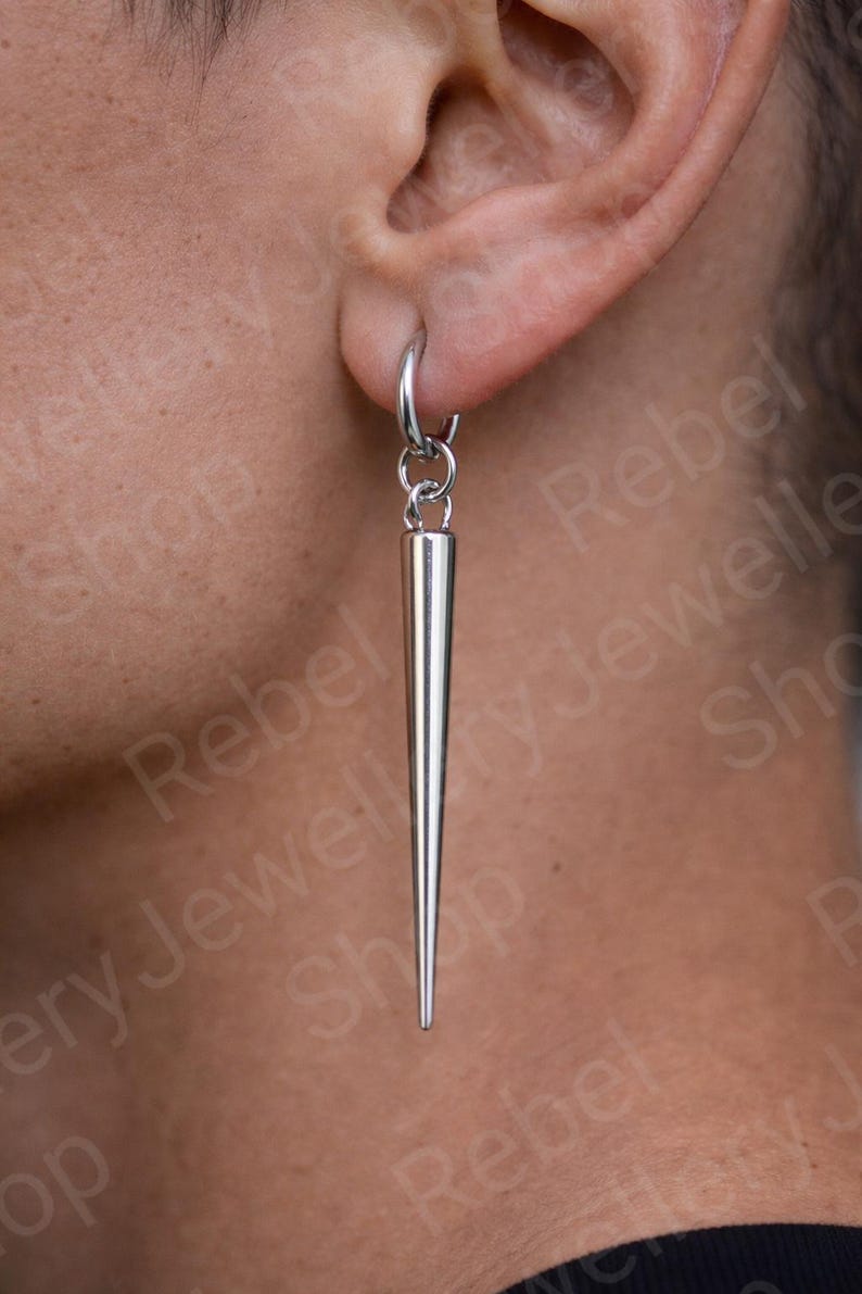 May include: A silver-colored, pointed earring is suspended from a small hoop. The earring has a long, conical shape, tapering to a sharp point. The jewelry is worn on the ear. The background is blurred.